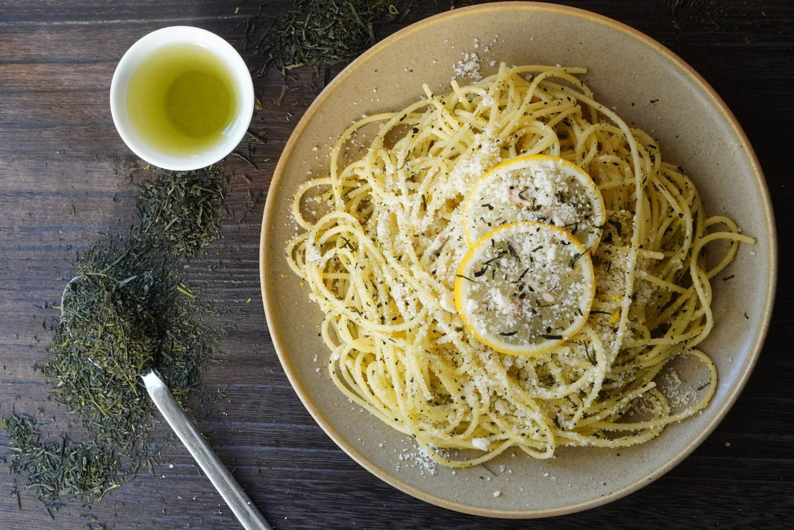 Plate of spaghetti with lemon slices and parmesan cheese, green tea , accompanied by a small bowl of olive oil on a wooden surface.
